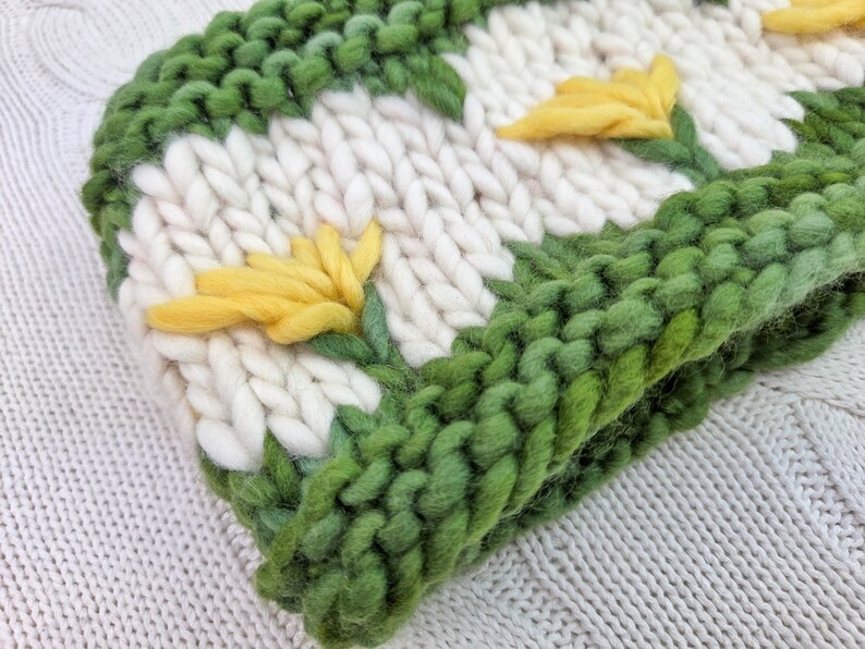 Puede incluir: Un gorro de punto grueso en crema, verde y amarillo. El gorro presenta un patr&oacute;n repetido de flores amarillas con tallos y hojas verdes sobre un fondo crema. El borde y los contornos de las flores son verdes.