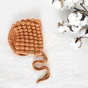 May include: A brown crocheted baby bonnet with a brown leather tag that says "Seach.ly". The bonnet has a long tie that can be tied under the chin.