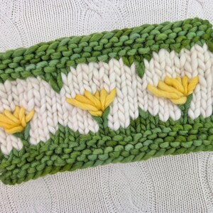 Puede incluir: Diadema tejida a mano con dise&ntilde;o de margaritas. La diadema es principalmente blanca con detalles verdes y amarillos. Las margaritas son amarillas con p&eacute;talos blancos, sobre un fondo verde. La diadema mide aproximadamente 10 cm de ancho.