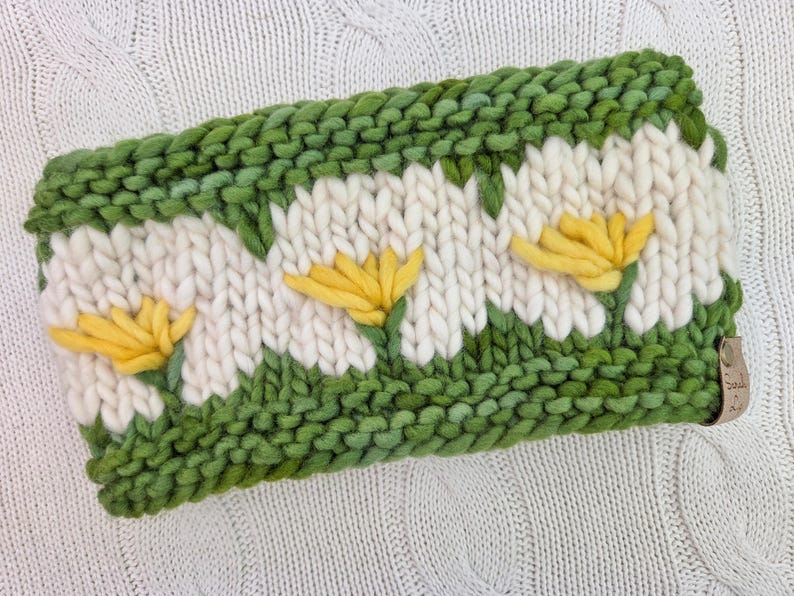Puede incluir: Diadema tejida a mano con dise&ntilde;o de margaritas. La diadema es principalmente blanca con detalles verdes y amarillos. Las margaritas son amarillas con p&eacute;talos blancos, sobre un fondo verde. La diadema mide aproximadamente 10 cm de ancho.