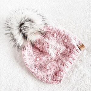 May include: A pink knitted beanie with a white and black faux fur pom pom. The beanie has a textured pattern with small raised dots.