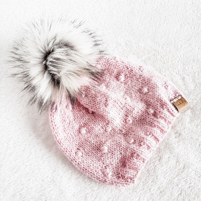 May include: A pink knitted beanie with a white and black faux fur pom pom. The beanie has a textured pattern with small raised dots.