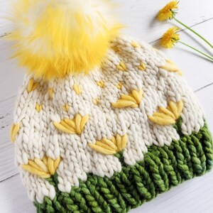 May include: White knitted beanie with a yellow pom pom and yellow flower accents. The beanie has a green brim.