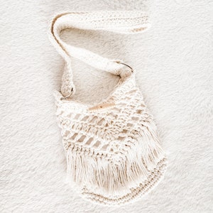 May include: A white crocheted bag with a long strap and fringe detailing. The bag has a wooden toggle closure.
