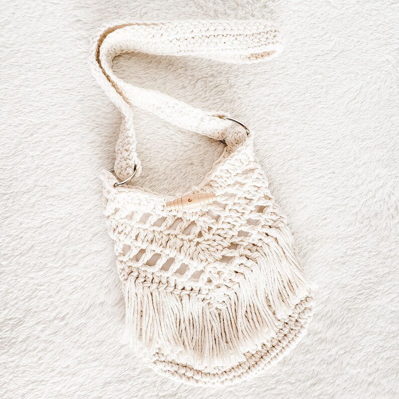 May include: A white crocheted bag with a long strap and fringe detailing. The bag has a wooden toggle closure.