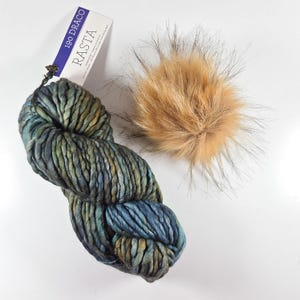 May include: A skein of thick, variegated yarn in shades of blue, green, and brown with a white paper tag labeled 190 DRACO RASTA, paired with a fluffy, round, light brown faux fur pom-pom featuring long, dark-tipped fibers for texture.