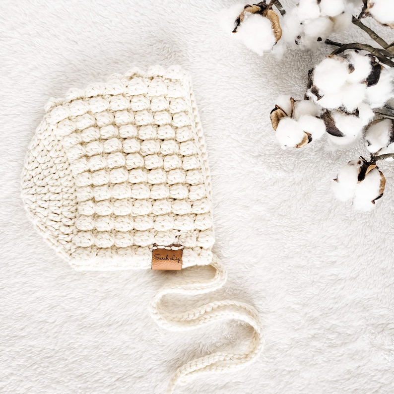 May include: A white crocheted baby bonnet with a brown leather tag that says "Sunkissed". The bonnet has a long tie that can be tied under the chin.
