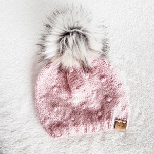 May include: A pink knitted beanie with a white pom pom. The beanie has a textured pattern with small white dots.