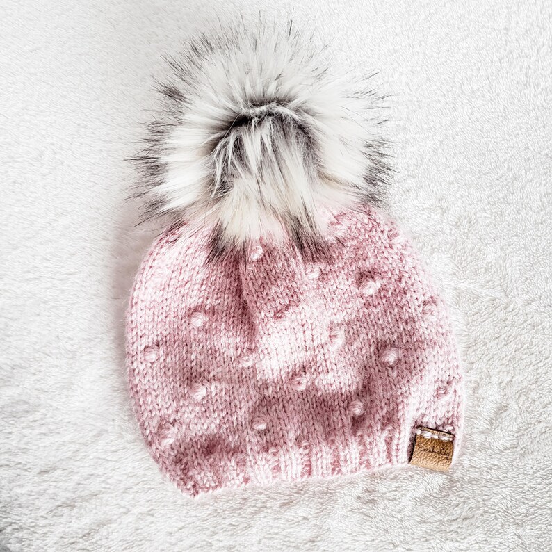 May include: A pink knitted beanie with a white pom pom. The beanie has a textured pattern with small white dots.