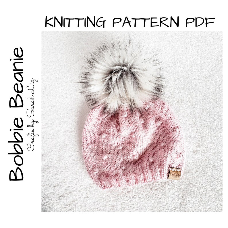 May include: A pink knitted beanie with a white and black faux fur pom pom. The beanie has a textured stitch pattern and a small brown leather tag.
