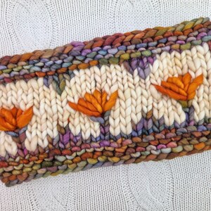 Puede incluir: Diadema tejida a mano con una base color crema y tres aplicaciones de flores naranjas. Los bordes son una mezcla de hilos coloridos, incluyendo tonos marrones, morados y verdes. Una peque&ntilde;a etiqueta de cuero con texto est&aacute; adjunta al costado.