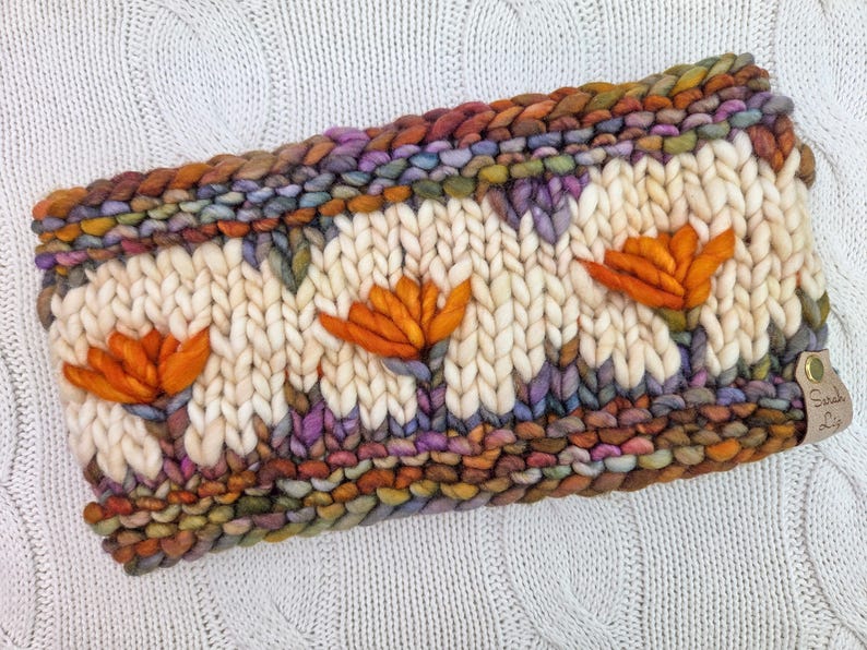 Puede incluir: Diadema tejida a mano con una base color crema y tres aplicaciones de flores naranjas. Los bordes son una mezcla de hilos coloridos, incluyendo tonos marrones, morados y verdes. Una peque&ntilde;a etiqueta de cuero con texto est&aacute; adjunta al costado.