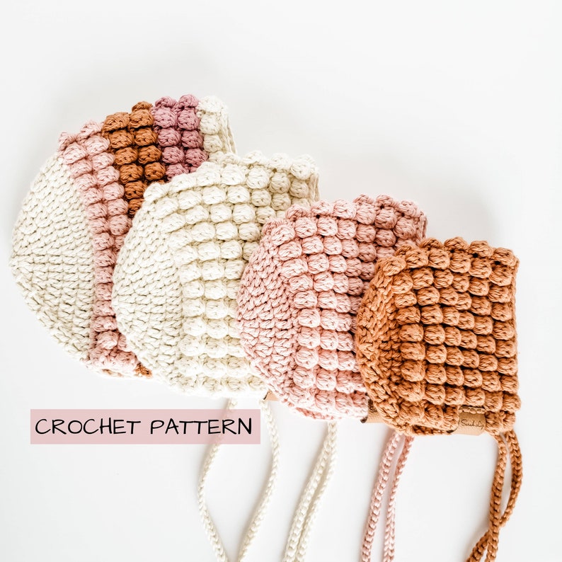 May include: Four crocheted hats in different shades of pink, beige, and brown. The hats have a textured, puffy design and are tied with white cords. The text "CROCHET PATTERN" is visible at the bottom of the image.