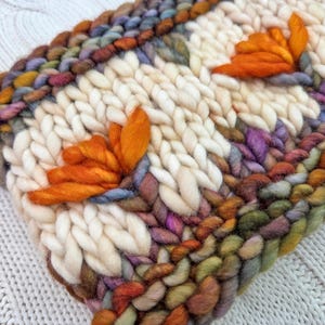 Puede incluir: Primer plano de un textil tejido a mano con un fondo crema y acentos florales naranjas. Los bordes est&aacute;n bordeados con hilos coloridos en tonos de morado, verde y marr&oacute;n. La textura es gruesa y acogedora.