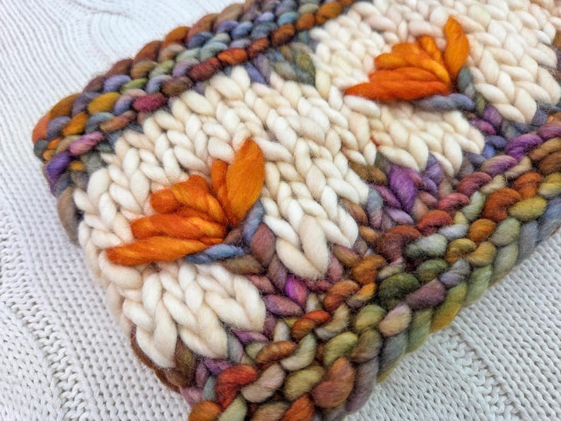 Puede incluir: Primer plano de un textil tejido a mano con un fondo crema y acentos florales naranjas. Los bordes est&aacute;n bordeados con hilos coloridos en tonos de morado, verde y marr&oacute;n. La textura es gruesa y acogedora.