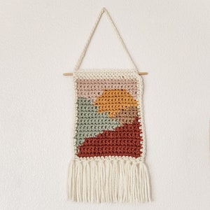 May include: A crocheted wall hanging with a geometric design in shades of pink, green, orange, and brown. The hanging has a white fringe at the bottom.