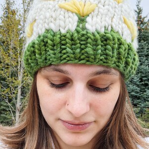 May include: A woman wearing a white and green knitted hat with a yellow pom-pom. The hat features a daisy design. She is holding a yellow dandelion.