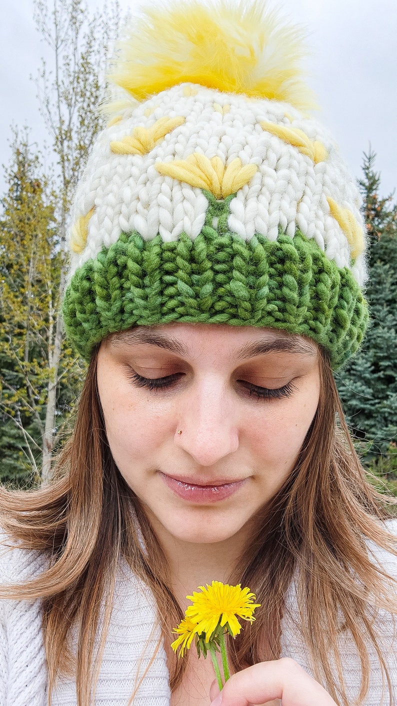 May include: A woman wearing a white and green knitted hat with a yellow pom-pom. The hat features a daisy design. She is holding a yellow dandelion.