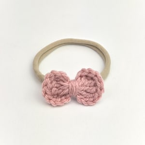 Op de afbeelding: Een lichtroze gehaakte strik-haarband. De strik is gecentreerd op een elastische band in neutrale kleur. De strik heeft een textuur en de haarband is geschikt voor baby's en jonge kinderen.