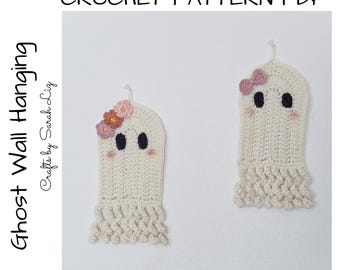 May include: Two handmade crochet ghost wall hangings. The ghosts are off-white with black eyes and pink blush. One ghost has a pink bow, the other has flowers. The text says "Ghost Wall Hanging" and "Crochet Pattern PDF".