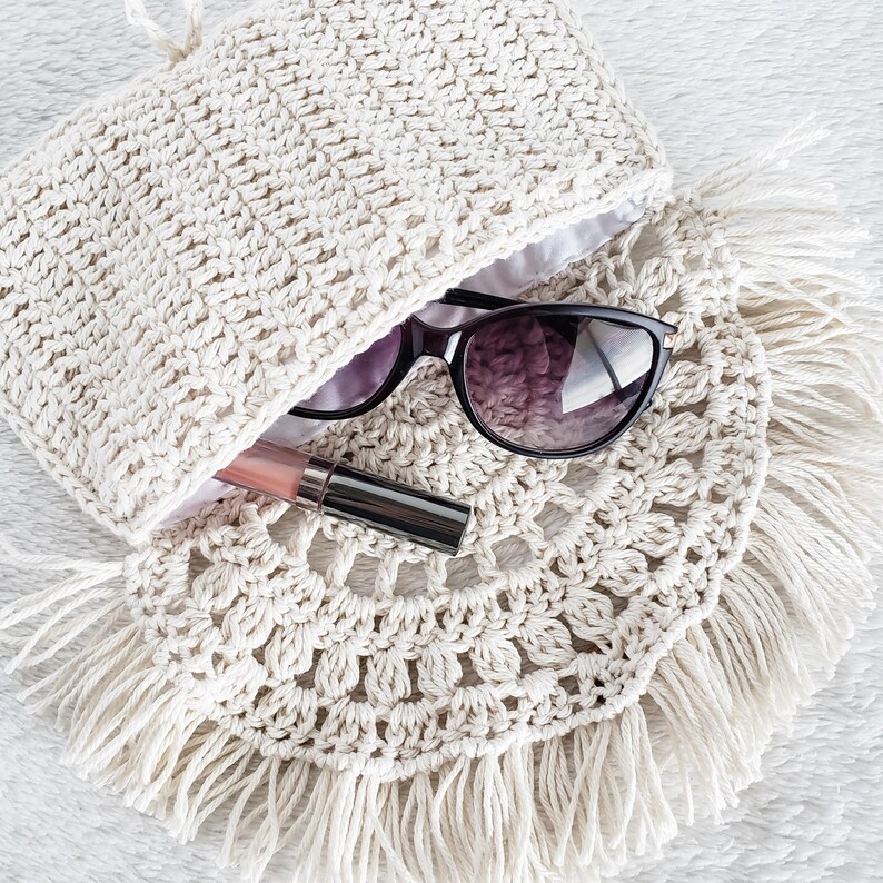 May include: A white crocheted bag with fringe detailing. The bag is open and a pair of sunglasses and a pink lip gloss are inside.