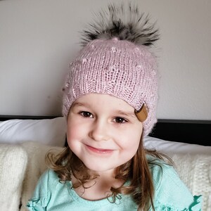 May include: A young girl wearing a pink knitted hat with a black pom pom and a brown leather patch. The hat has a textured design and is pulled down over her ears. She is smiling and looking at the camera.