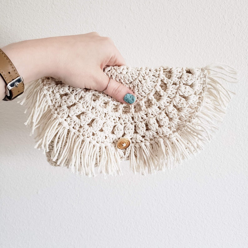 May include: A crocheted clutch bag with a fringe trim and a button closure. The bag is made of a cream-colored yarn and has a natural, bohemian style.