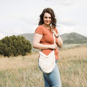 May include: A woman wearing a rust-coloured t-shirt and blue jeans is standing in a field. She is holding a white crocheted bag with fringe.