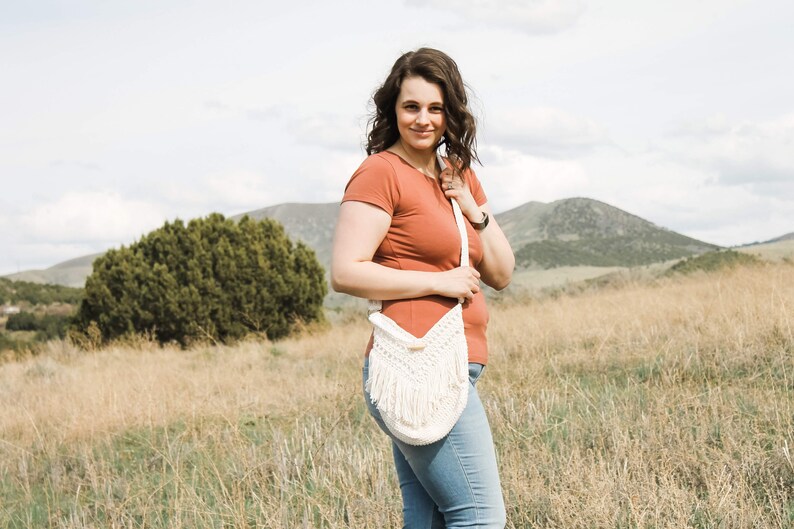May include: A woman wearing a rust-coloured t-shirt and blue jeans is standing in a field. She is holding a white crocheted bag with fringe.