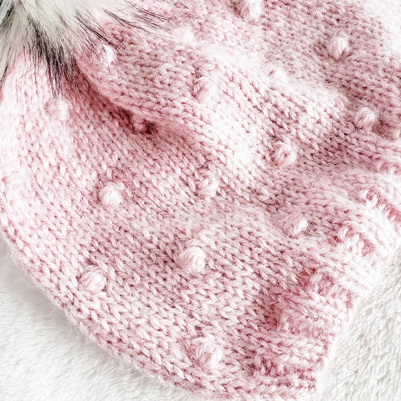 May include: A close-up of a pink knitted hat with a textured bobble stitch. The hat has a fluffy white pom-pom on top.