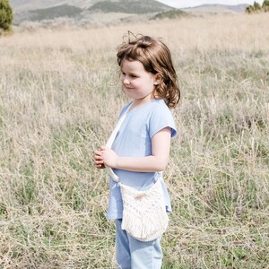 May include: A young girl wearing a light blue shirt and blue trousers stands in a field of long grass. She is holding a white crocheted bag with a fringe.