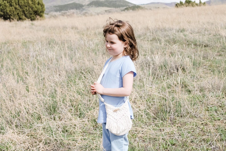 May include: A young girl wearing a light blue shirt and blue trousers stands in a field of long grass. She is holding a white crocheted bag with a fringe.
