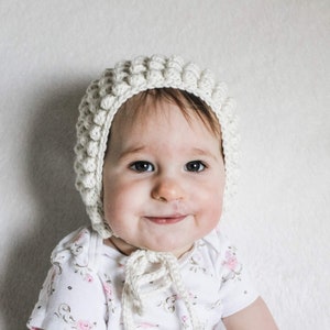 May include: A baby wearing a white crocheted bonnet with a floral pattern on their dress.