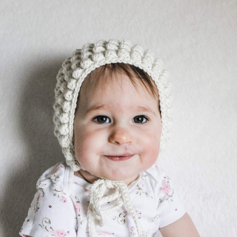 May include: A baby wearing a white crocheted bonnet with a floral pattern on their dress.