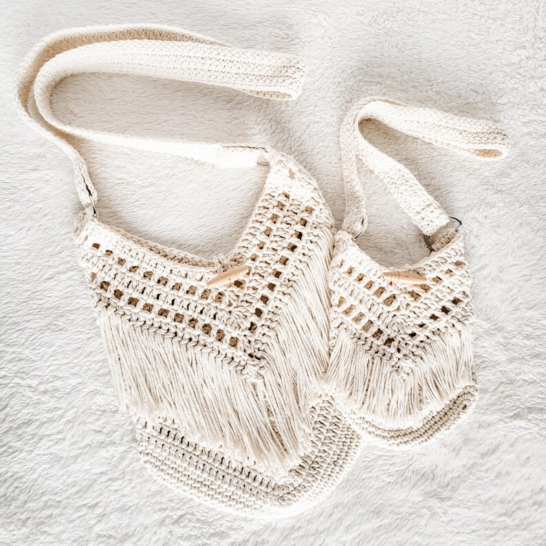 May include: Two crocheted bags with fringe detailing. The larger bag has a long strap, while the smaller bag has a shorter strap. Both bags are made of natural white yarn.