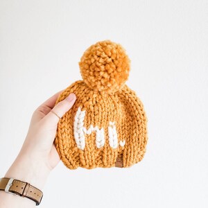 以下が含まれることがあります： 白いポンポンと白い刺繍で「こんにちは」と書かれたマスタードイエローのニットビーニー
