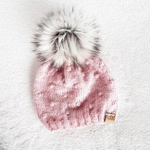 May include: A pink knitted beanie with a white pom pom. The beanie has a textured pattern with small raised dots.