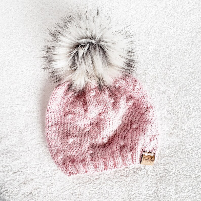 May include: A pink knitted beanie with a white pom pom. The beanie has a textured pattern with small raised dots.