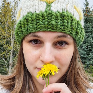 May include: A woman wearing a white and green knit hat with a yellow pom pom holds a yellow dandelion flower to her nose.