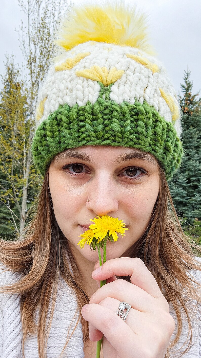 May include: A woman wearing a white and green knit hat with a yellow pom pom holds a yellow dandelion flower to her nose.