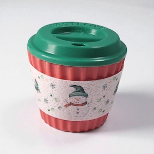 May include: A red and green reusable coffee cup with a festive winter design. The cup has a red, ribbed body and a green lid. A white band with a snowman and snowflake pattern wraps around the cup.