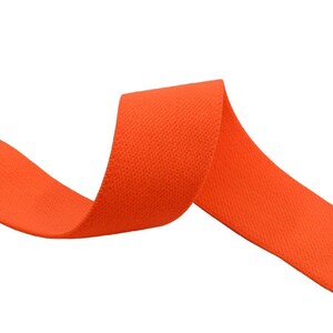 K&ouml;nnte beinhalten: Orangefarbenes Gummiband, eine Art Stoffbesatz, in einer gekr&auml;uselten Form. Das Band besteht aus einem gewebten Material mit einer strukturierten Oberfl&auml;che.