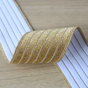 Könnte beinhalten: Gold- und weiß gestreiftes elastisches Band mit glitzerndem Finish. Das Band ist in der Mitte gefaltet und zeigt beide Seiten.