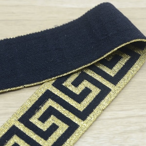 Może przedstawiać: Czarna elastyczna lamówka z złotym wzorem greckiego klucza. Lamówka ma szerokość około 2,5 cm.
