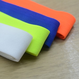 K&ouml;nnte beinhalten: Vier Rollen elastischen Stoffes in verschiedenen Farben: Wei&szlig;, Neongelb, Blau und Orange. Der Stoff ist gefaltet und auf einer Holzoberfl&auml;che gestapelt.