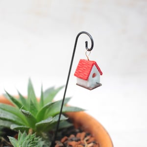 Könnte beinhalten: Ein Miniatur-Vogelhaus in Weiß mit rotem Dach hängt an einem schwarzen Metallhaken. Das Vogelhaus ist an einem schwarzen Metallstab befestigt, der in einen Terrakotta-Topf mit grünen Sukkulenten gesteckt wird.