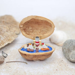 Peut inclure: Une scène miniature de paysage marin créée à l'intérieur d'une noix de noyer coupée en deux. La scène représente un phare, des voiliers et une petite île avec des palmiers, le tout réalisé en argile colorée.