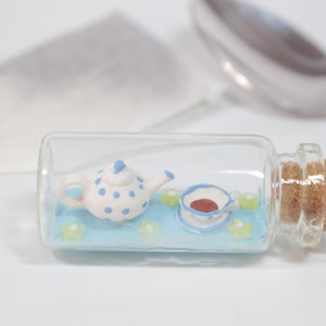 Könnte beinhalten: Eine kleine Glasflasche mit einem Korkverschluss enthält eine Miniatur-Teekanne in Weiß mit blauen Punkten und eine passende Teetasse und Untertasse. Die Teekanne und die Teetasse befinden sich auf einer blauen Oberfläche mit grünen Blumen.