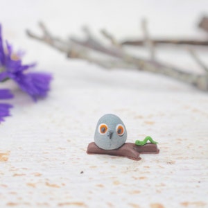 May include: A small gray owl figurine with orange eyes perched on a brown branch with a green leaf. The owl is made of clay and has a whimsical look.