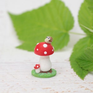 Könnte beinhalten: Eine skurrile Miniatur-Pilzfigur mit rotem Hut und weißen Punkten, einem weißen Stiel und einer kleinen Schnecke obenauf. Ein kleinerer Pilz sitzt am Fuß, alles auf einer grünen Basis. Der Hintergrund ist unscharf.